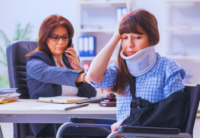 An injured person sitting in a wheelchair, wearing a neck brace and an arm sling, holding their head with one hand. Across the desk, a professional is engaged in a phone call, likely discussing legal or insurance matters related to the injury.