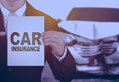 A businessman holding a sign that reads "CAR INSURANCE" with a damaged car in the background, highlighting the concept of car insurance as protection against vehicle damage.