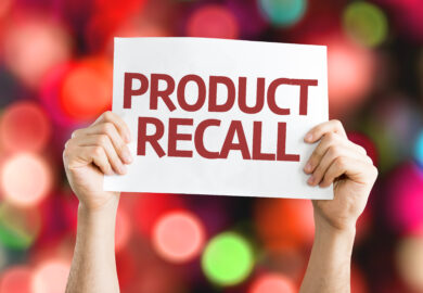 Hands holding product recall printed on a white paper with blurry lights in the background.