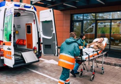 Paramedics assisting a patient on a stretcher into an ambulance outside a medical facility.