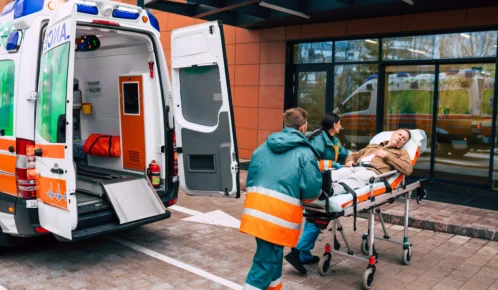 Paramedics assisting a patient on a stretcher into an ambulance outside a medical facility.