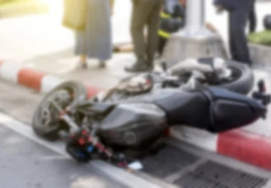 Blurred image of a motorcycle lying on its side after an accident, with people and emergency responders in the background.