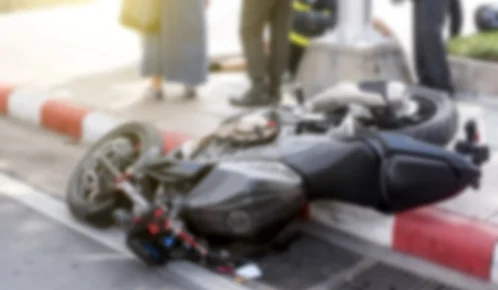 Blurred image of a motorcycle lying on its side after an accident, with people and emergency responders in the background.