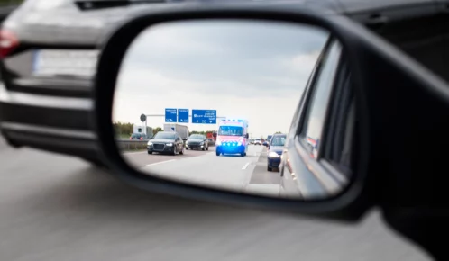 Ambulance with flashing lights seen in a car’s side mirror on a highway.