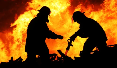 Two firefighters silhouetted against intense flames, working together to control a large blaze.