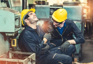 Two factory workers in safety gear, one sitting on the floor in pain holding his arm, while the other kneels beside him offering help.