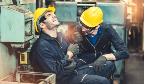 Two factory workers in safety gear, one sitting on the floor in pain holding his arm, while the other kneels beside him offering help.