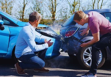 Two men crouched near their cars after a traffic accident. One man is by the front of a blue car with visible damage, and the other is near the rear of a gray car, which also has noticeable damage. Driver Lies About a Car Accident