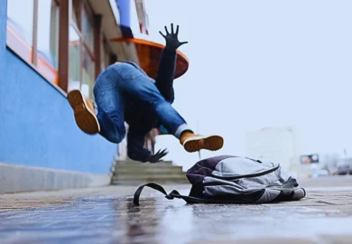 A person tripping and falling forward on a sidewalk after catching their foot on a backpack lying on the ground. Sidewalks Become Slip & Fall Traps