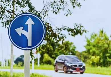 A blue circular traffic sign with two white arrows. One arrow points straight ahead, and the other curves left, indicating that drivers in this lane can either go straight or turn left. Who’s Liable in a Left-Turn Car Crash