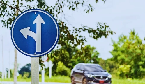 A blue circular traffic sign with two white arrows. One arrow points straight ahead, and the other curves left, indicating that drivers in this lane can either go straight or turn left. Who’s Liable in a Left-Turn Car Crash
