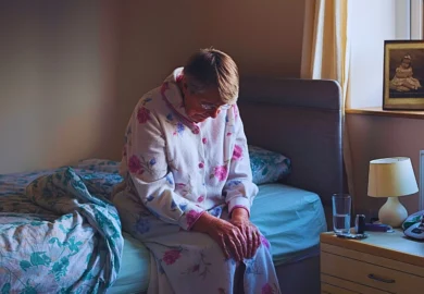 An elderly person sitting on the edge of a bed in a softly lit room.