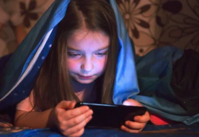 Young girl using a smartphone under a blue blanket. Social Media Platforms Enable Child Grooming