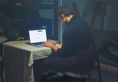 Man in dark clothing using laptop in dimly lit room. sue for sextortion