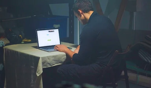 Man in dark clothing using laptop in dimly lit room. sue for sextortion