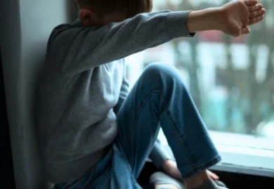 Sad and ashamed kid sitting in front a window with his arms hidding his face