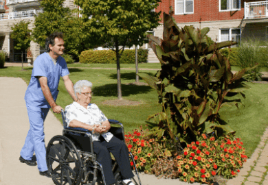 A male nurse wheels an elderly woman through a neighborhood.